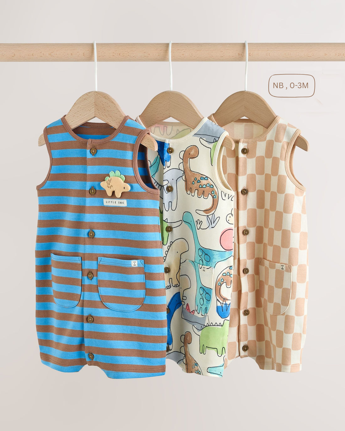 Three baby boys' sleeveless rompers hanging on wooden hangers — blue and brown stripe with dinosaur appliqué, cream all-over dinosaur print, and beige checkerboard design.