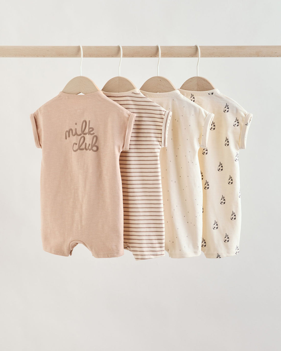 Front view of four baby rompers hanging on wooden hangers, showing the snap-front openings and short sleeves in a soft neutral colour palette.