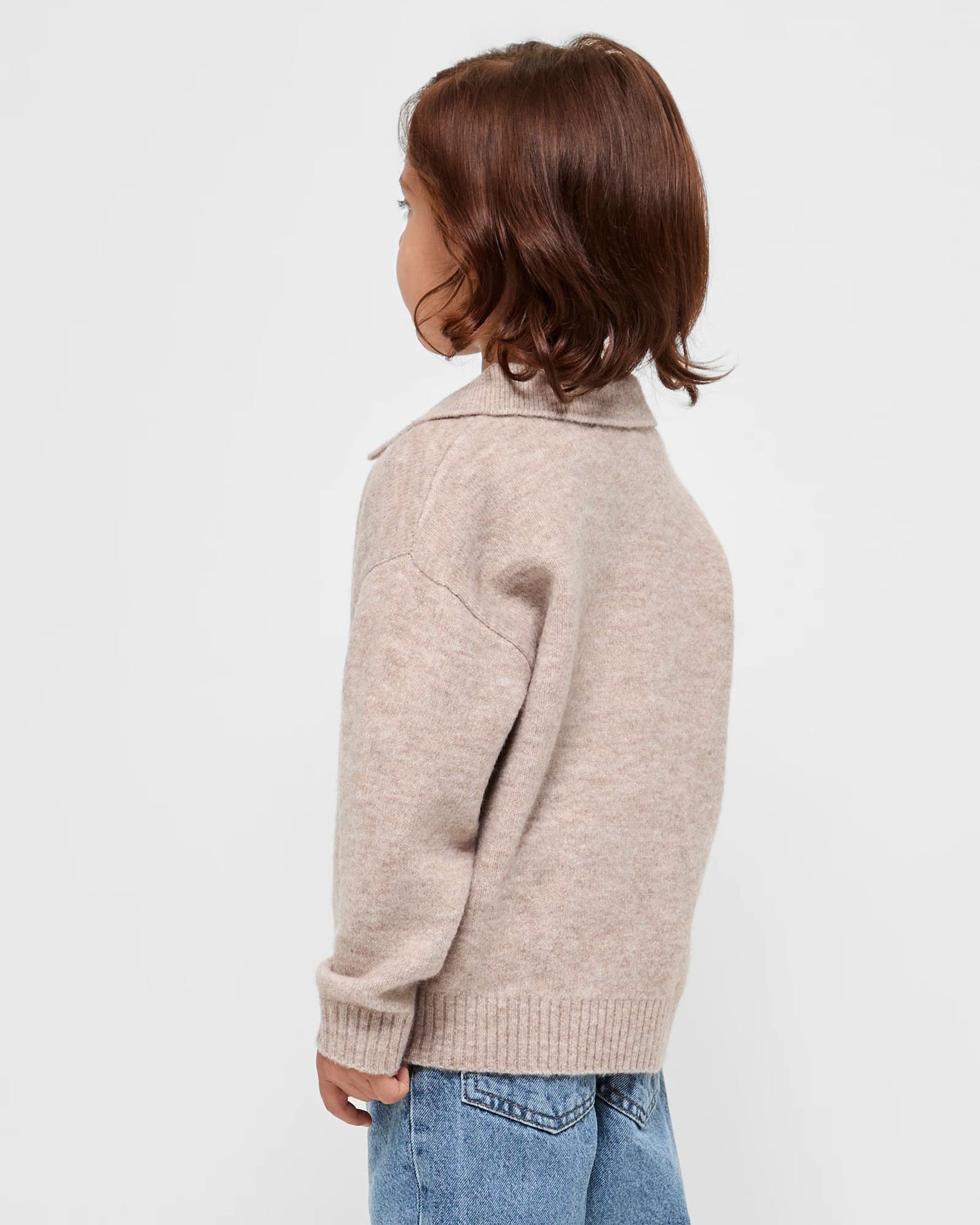 A kid wearing a beige sweater and blue jeans on a light gray background