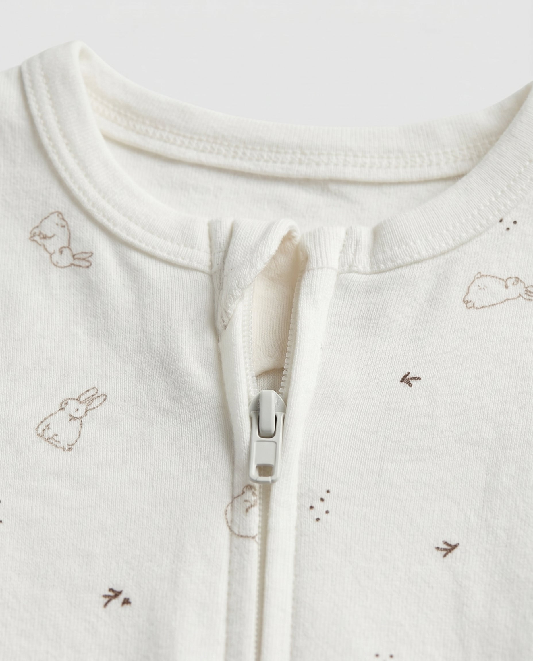 Ivory baby long-sleeve zip-up romper with all-over scattered bunny and bird print in taupe, featuring front zip closure with inner guard flap and ribbed cuffs at wrists and ankles