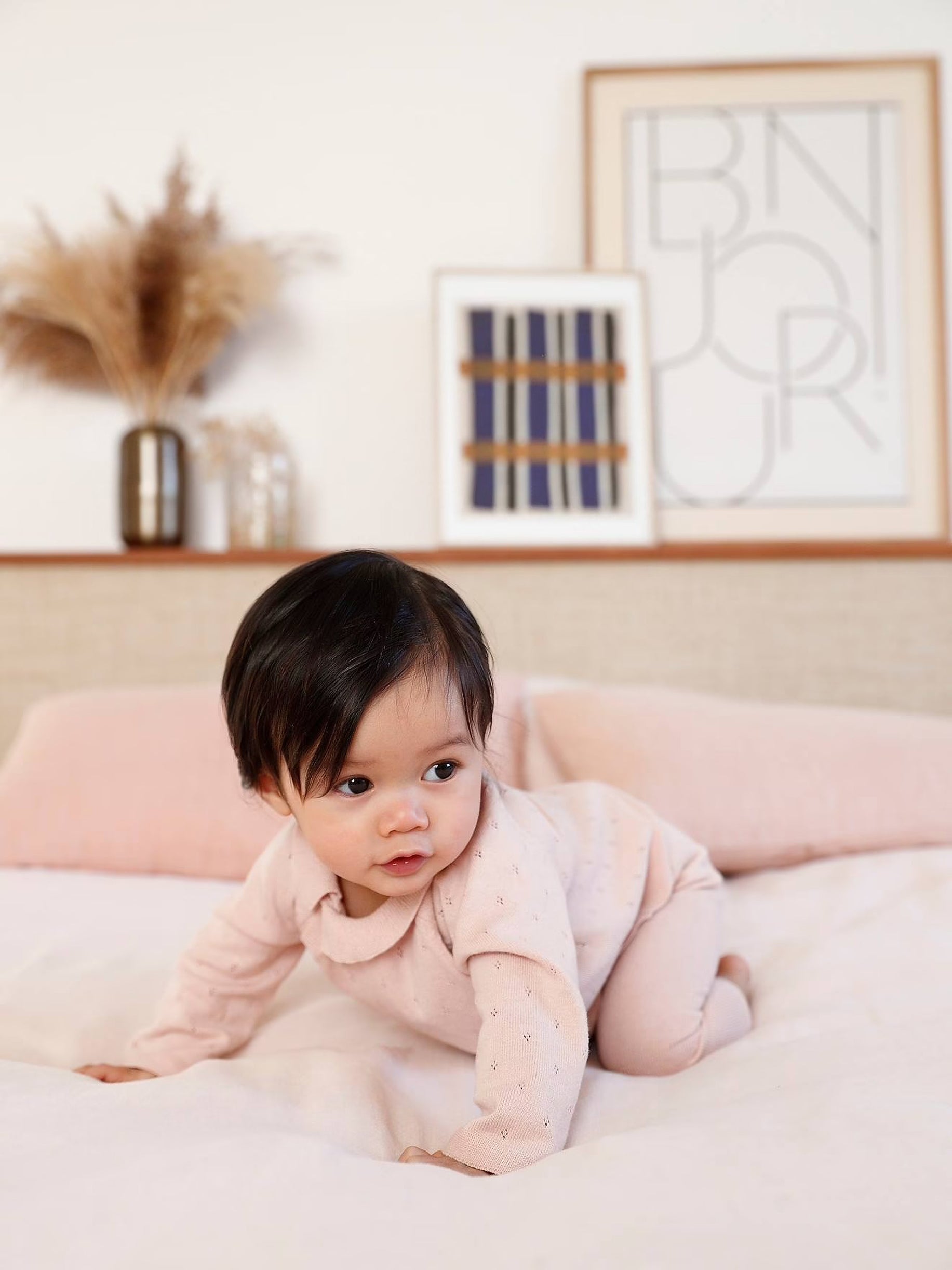 Baby in a pink outfit lying on a white blanket with a neutral-colored wall and decorative elements in the background.