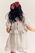 A child viewed from behind wearing a light beige linen dress with puff sleeves, a ruffled waist, and a large dusty rose bow in their dark hair, set against a plain off-white background.