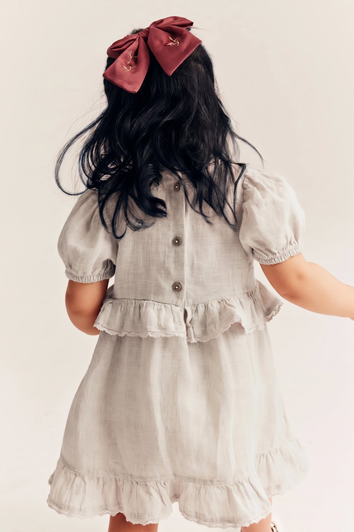 A child viewed from behind wearing a light beige linen dress with puff sleeves, a ruffled waist, and a large dusty rose bow in their dark hair, set against a plain off-white background.