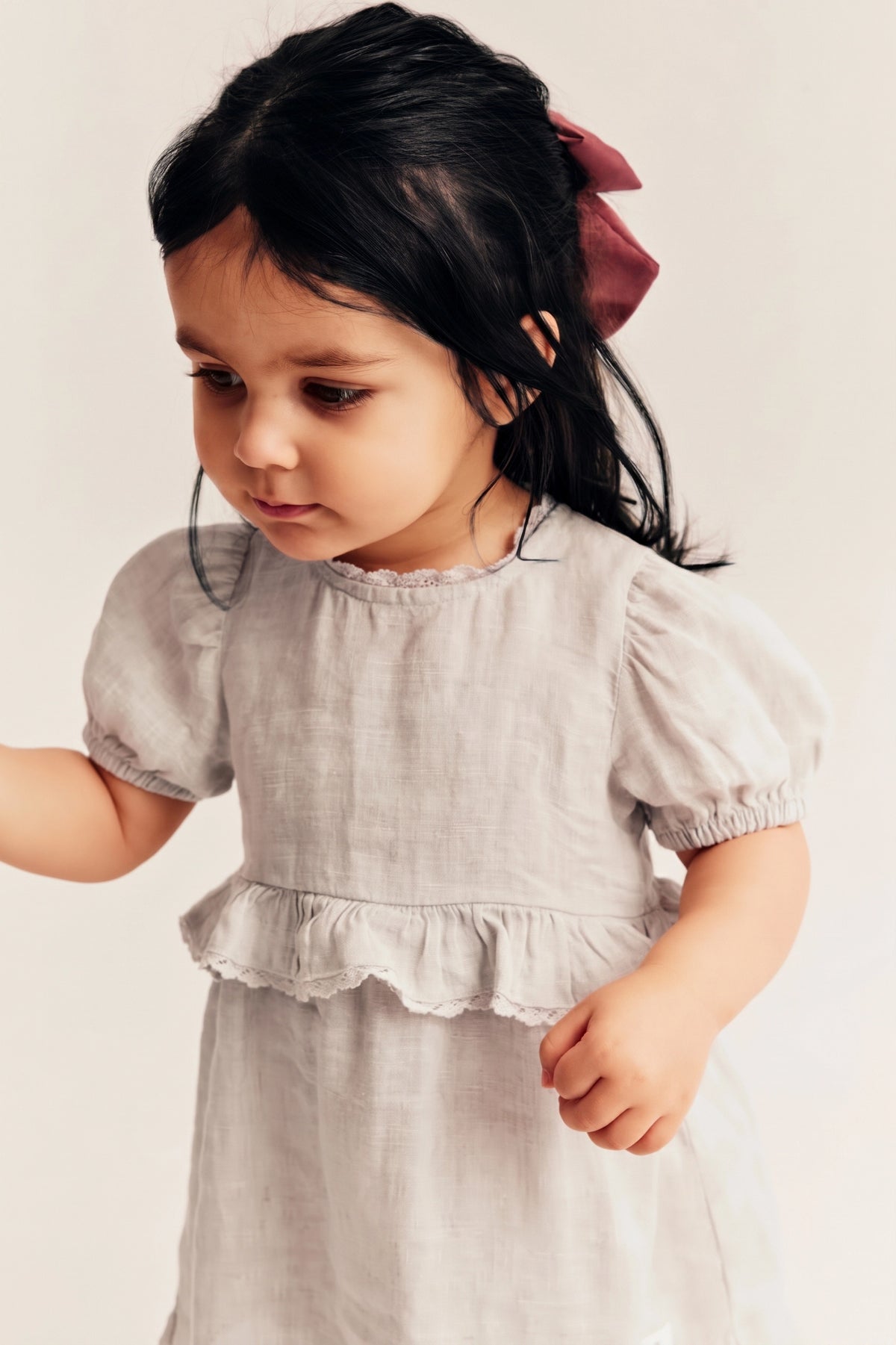 A toddler girl wearing a light beige linen dress with short puff sleeves and a ruffled hem, styled with a dusty rose hair bow, against a plain off-white background.