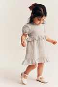 A toddler girl wearing a light gray linen dress with ruffled trim and white Mary Jane shoes, standing against a plain off-white background.