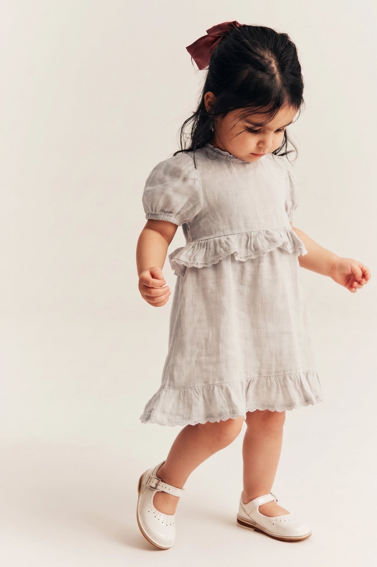 A toddler girl wearing a light gray linen dress with ruffled trim and white Mary Jane shoes, standing against a plain off-white background.