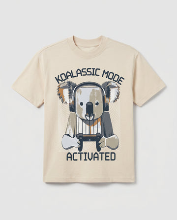 
Boys' cream-colored short-sleeve cotton t-shirt laid flat, featuring a graphic print of a cartoon koala wearing headphones and holding a gaming controller, with arched text reading "Koalassic Mode Activated" in dark navy.