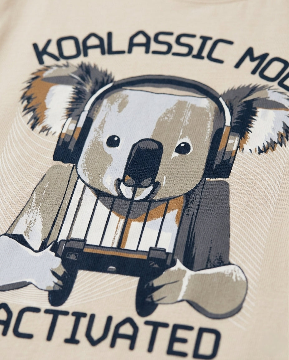 
Boys' cream-colored short-sleeve cotton t-shirt laid flat, featuring a graphic print of a cartoon koala wearing headphones and holding a gaming controller, with arched text reading "Koalassic Mode Activated" in dark navy.