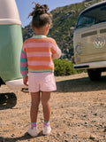 Child in a colorful striped sweater and pink shorts standing near a vintage Volkswagen van in a natural setting.
