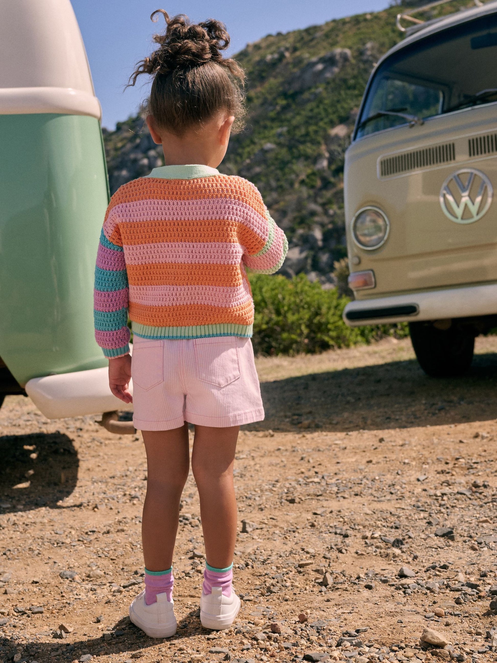 Child in a colorful striped sweater and pink shorts standing near a vintage Volkswagen van in a natural setting.