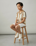Child sitting on a wooden stool wearing a striped shirt and white shorts.