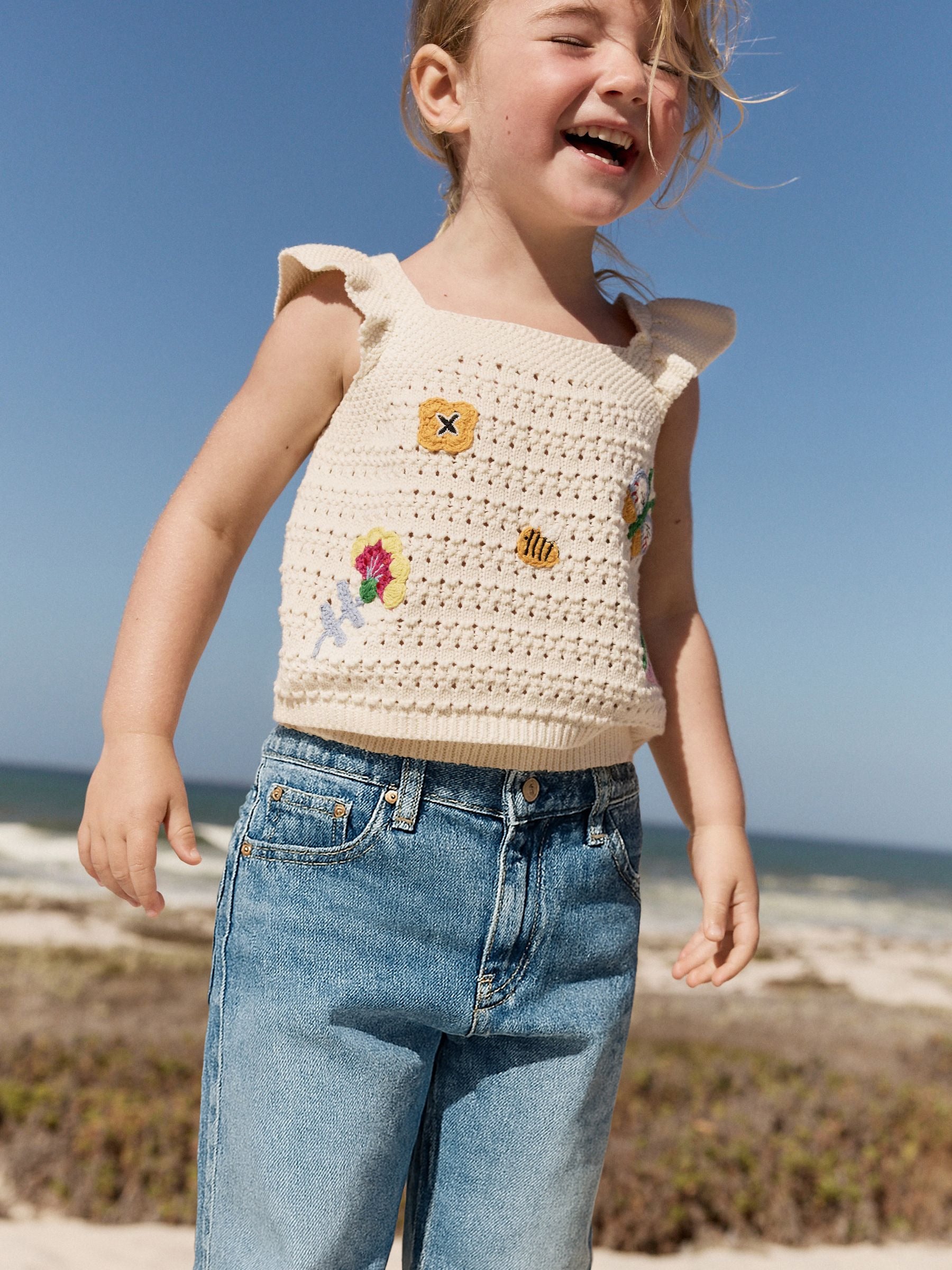 Child wearing a beige crochet top with floral and butterfly designs on a beach.