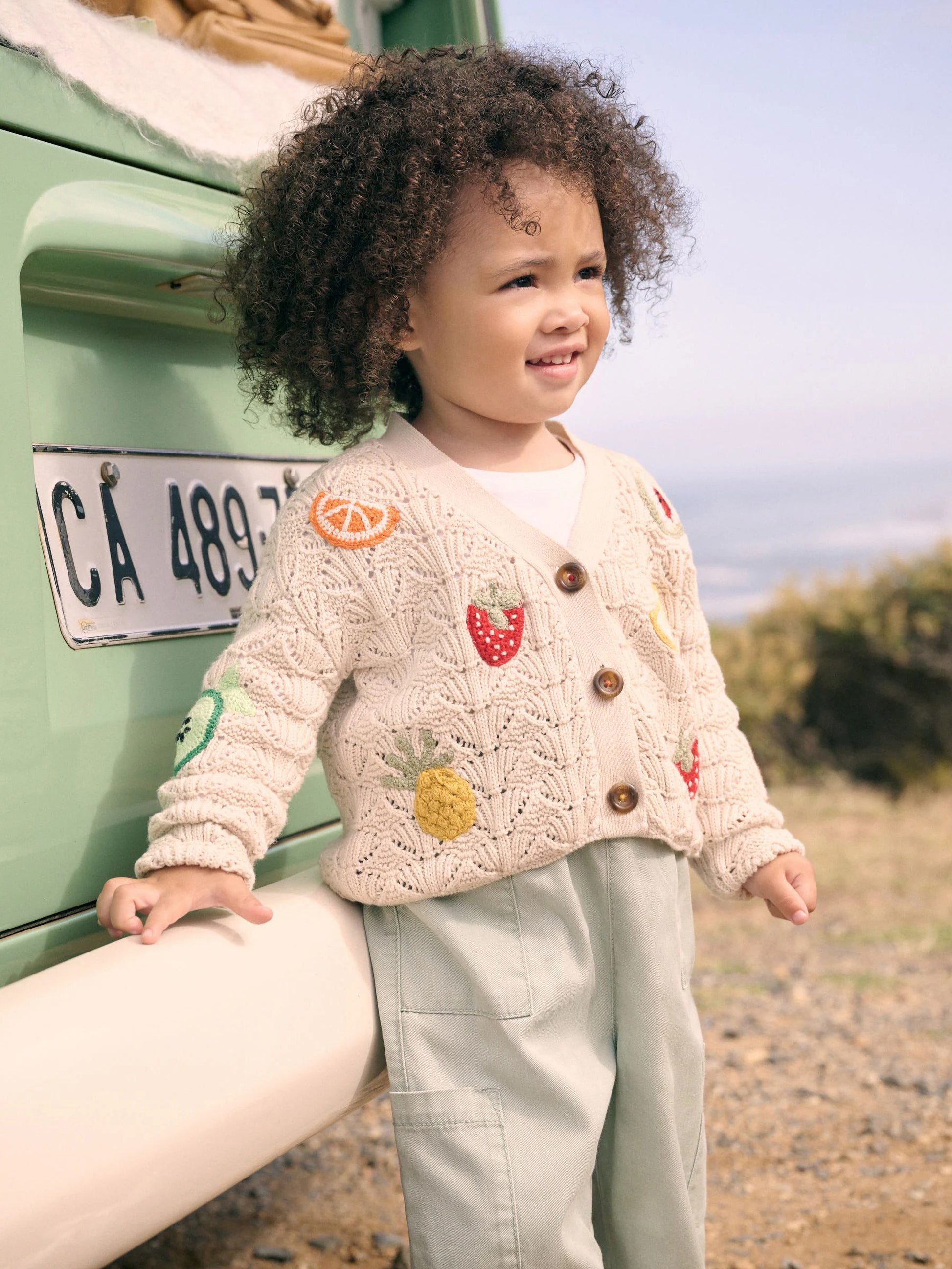 Child wearing a decorative sweater with buttons and patches, standing next to a vintage green truck.