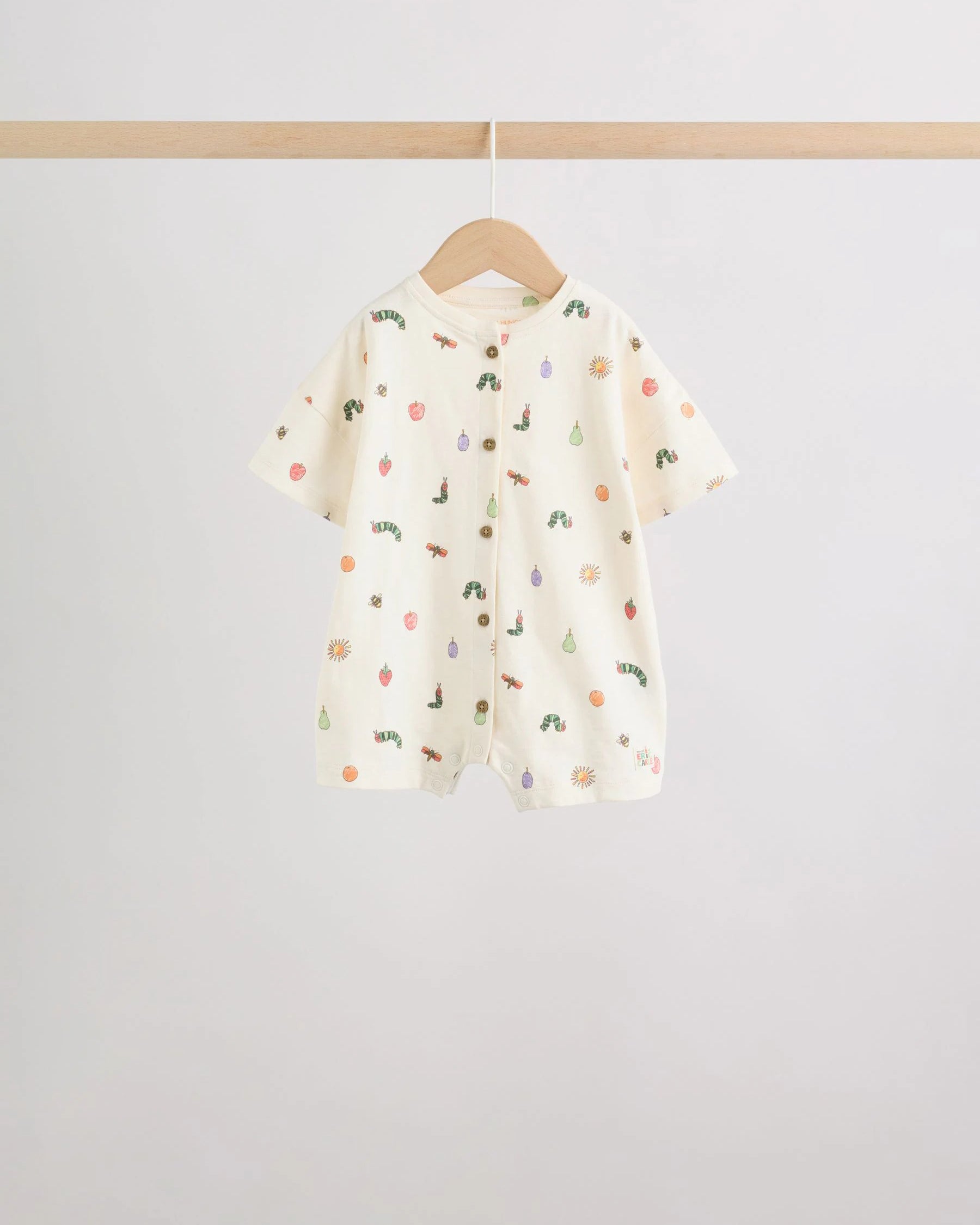 Cream all-over print baby romper with scattered caterpillar and fruit motifs, wooden button front placket, on wooden hanger

