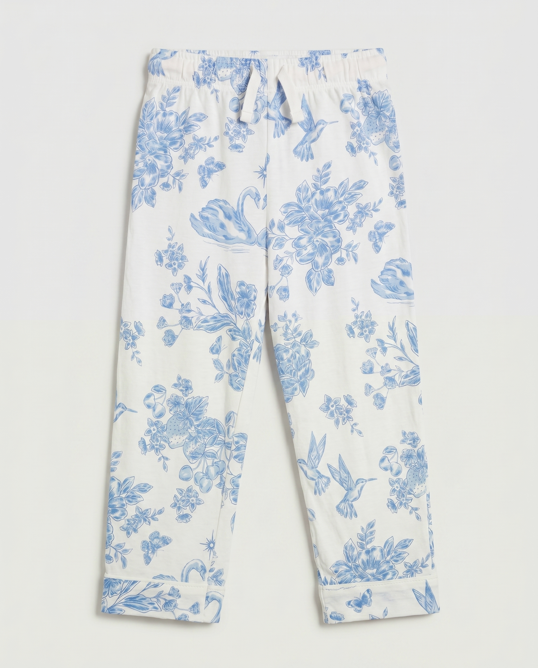 Full flat lay front view:
Girls' pyjama pants in ivory with an all-over blue toile de Jouy print featuring hummingbirds, swans, strawberries, and floral motifs, displayed flat lay on a white background with a relaxed straight leg and folded cuff hem.