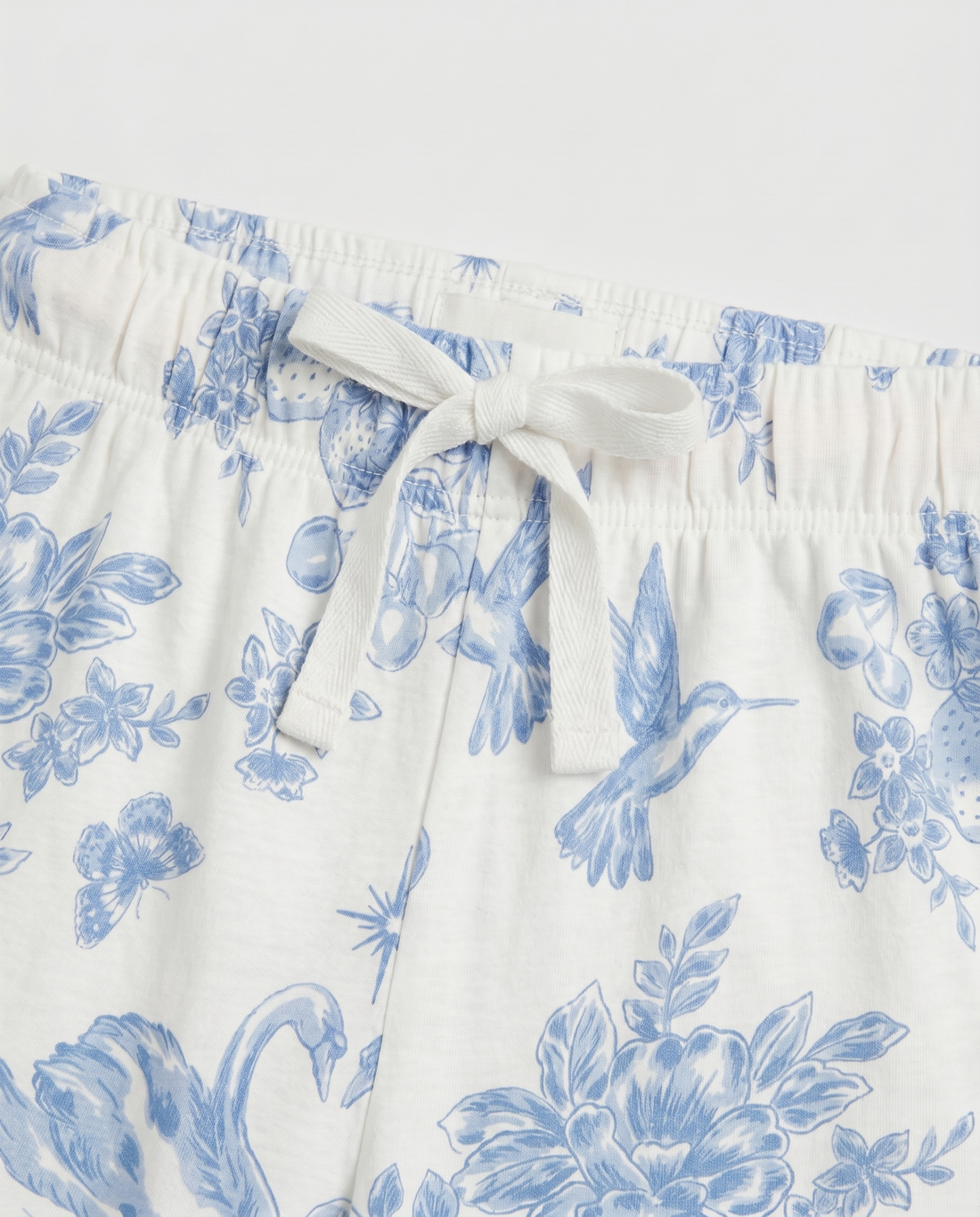 Full flat lay front view:
Girls' pyjama pants in ivory with an all-over blue toile de Jouy print featuring hummingbirds, swans, strawberries, and floral motifs, displayed flat lay on a white background with a relaxed straight leg and folded cuff hem.