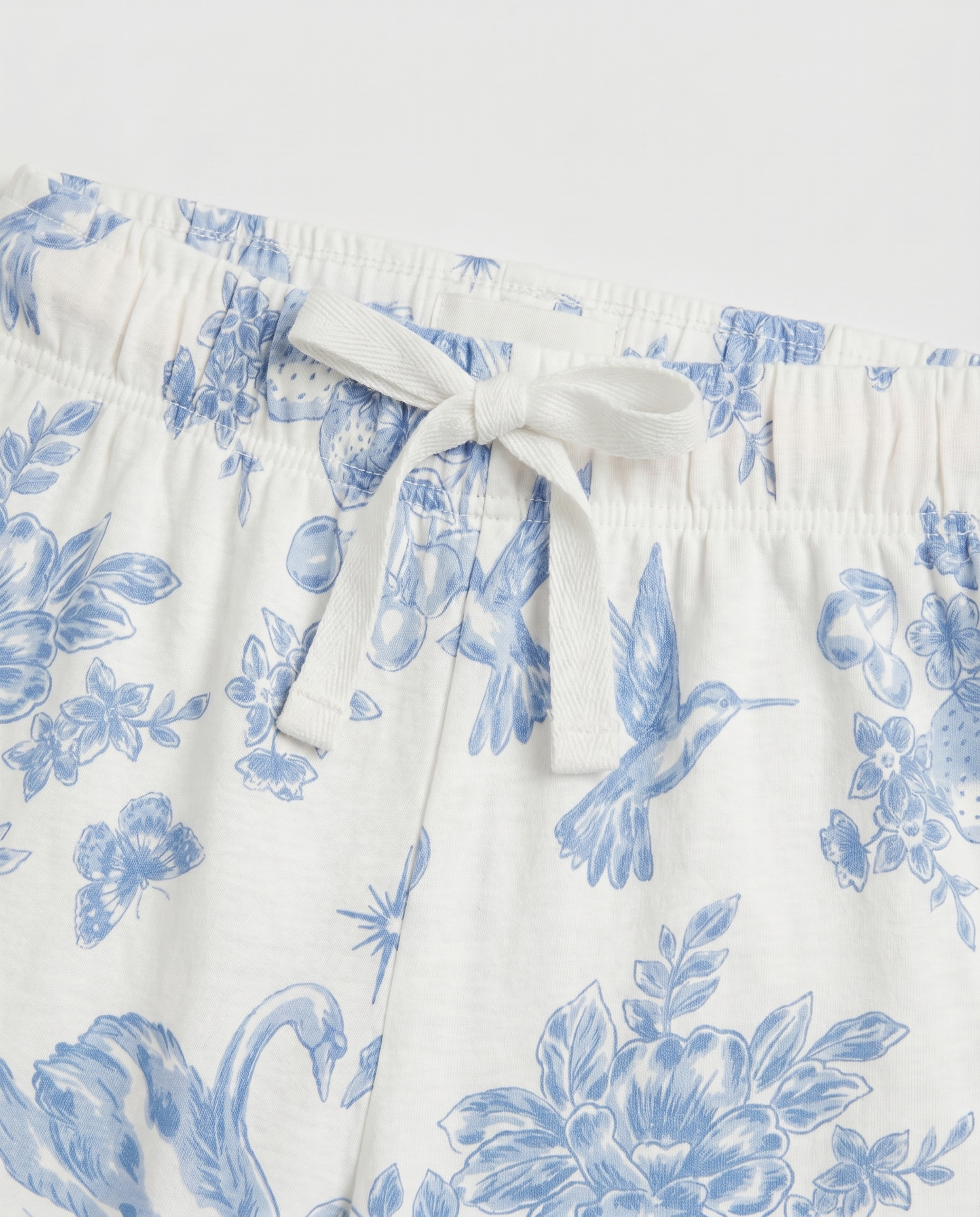 Close-up waistband detail:
Close-up of the elasticated waistband on girls' toile de Jouy pyjama pants, showing a woven ivory drawstring tied in a bow with blue floral and bird print fabric detail.