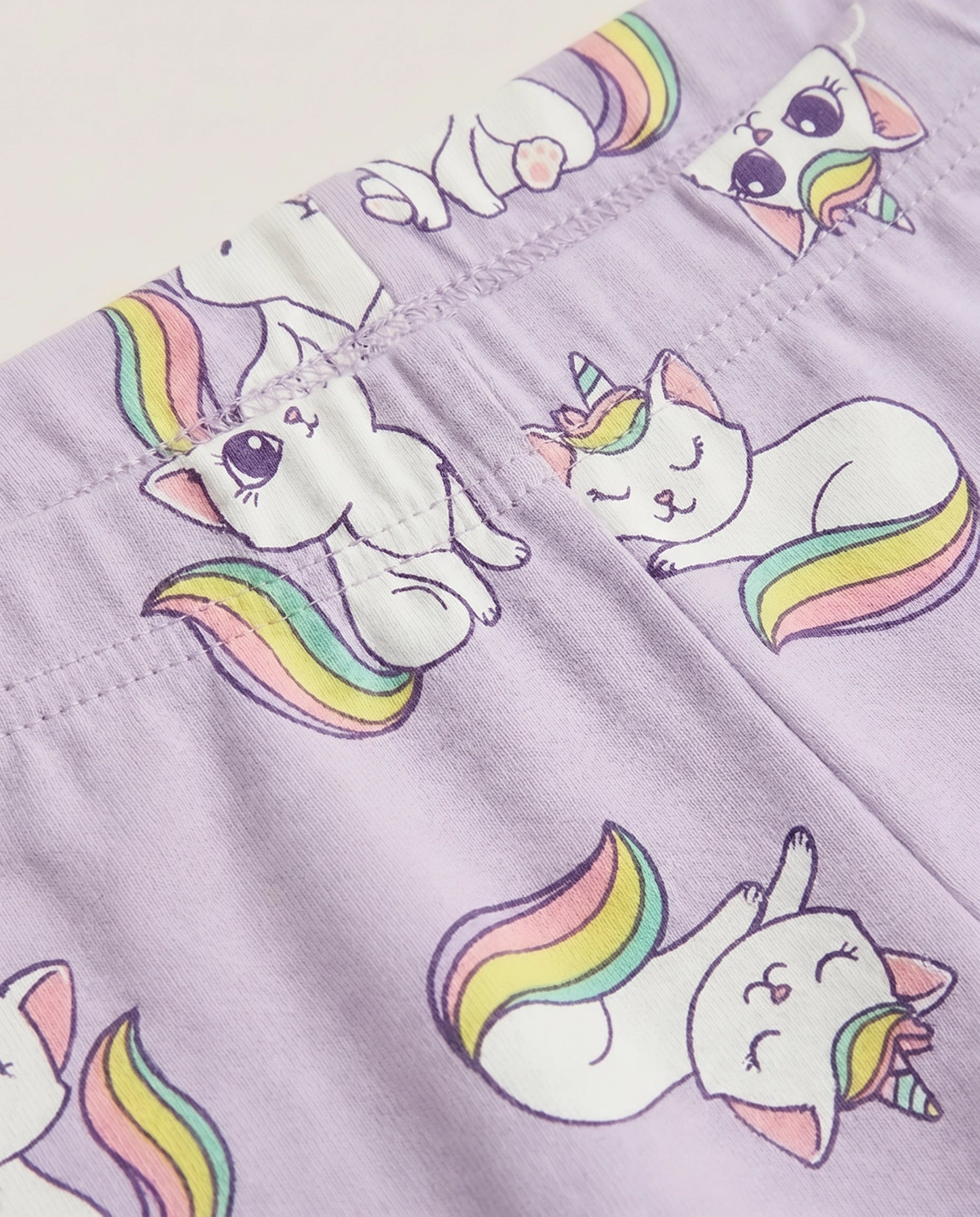 Close-up of lavender kids' leggings with all-over kawaii unicorn cat print featuring white cats with rainbow tails and unicorn horns on a purple background.