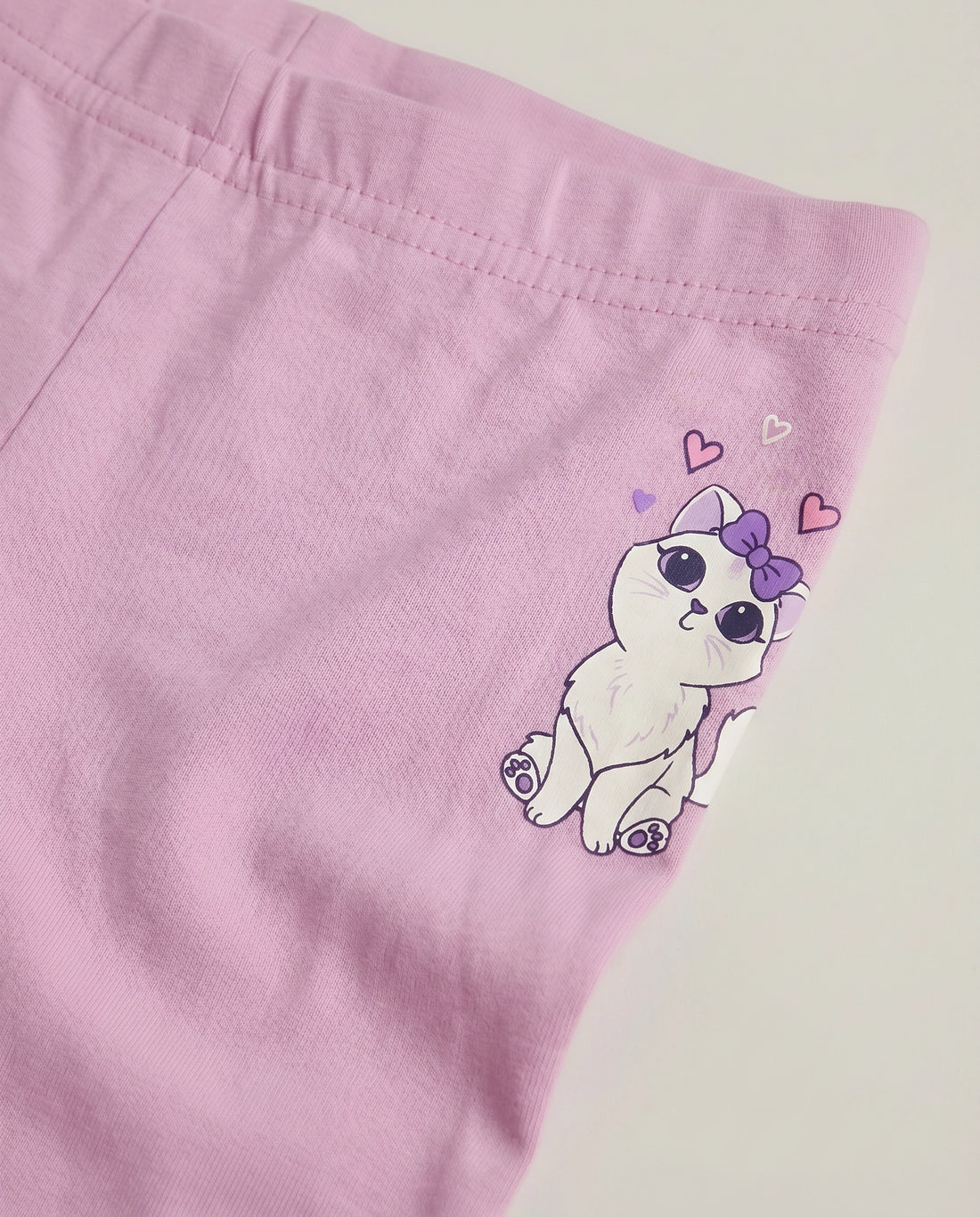 Close-up of girls' soft pink cotton leggings featuring a kawaii white cat graphic with a purple bow, floating hearts and gold stars at the hip, with elasticated waistband detail visible.