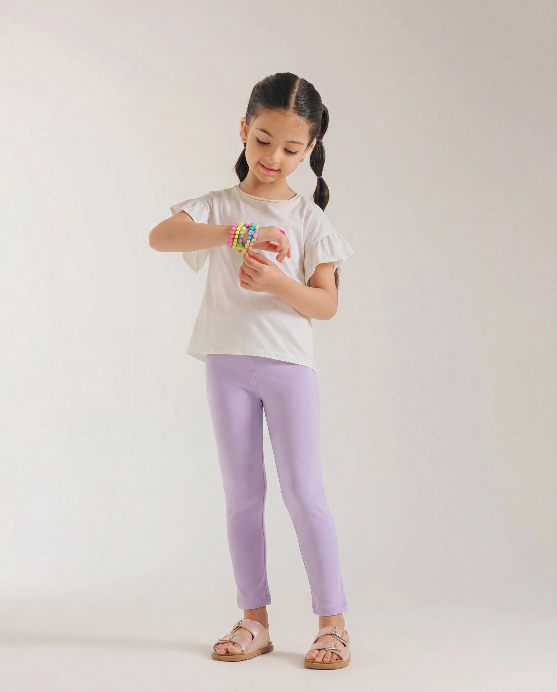 Young girl wearing baby purple full-length cotton leggings styled with a cream flutter-sleeve top and nude sandals, standing against a light grey background.