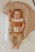 Baby in a brown and white striped shirt and beige pants holding a teddy bear, lying on a textured pillow.