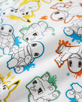 Close-up of a white kids' t-shirt fabric showing an all-over comic-style print of multiple Pokémon characters including Pikachu, Squirtle, Charmander, and Bulbasaur with colourful paint-splash backgrounds in yellow, blue, and orange