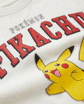 Close-up of an off-white kids' t-shirt showing bold red varsity-style Pikachu lettering and a yellow Pikachu character graphic with a playful open-mouth expression