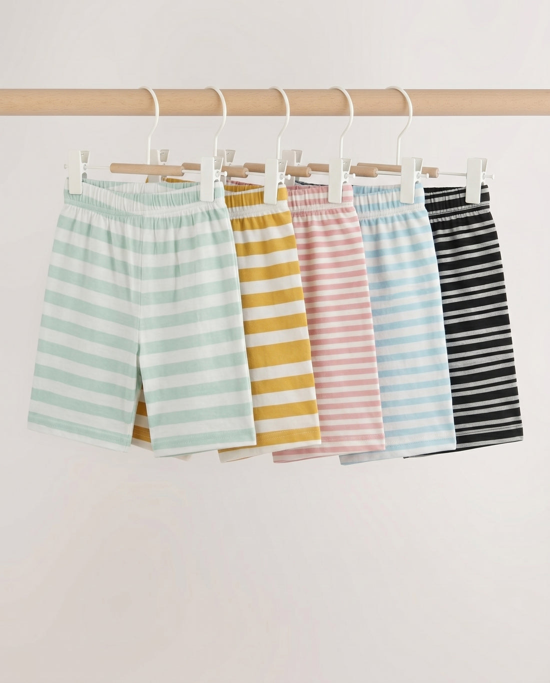 Five kids' striped pull-on shorts fanned out flat lay — mint, mustard, pink, blue, and black stripe patterns with elastic waistband, on light grey background.