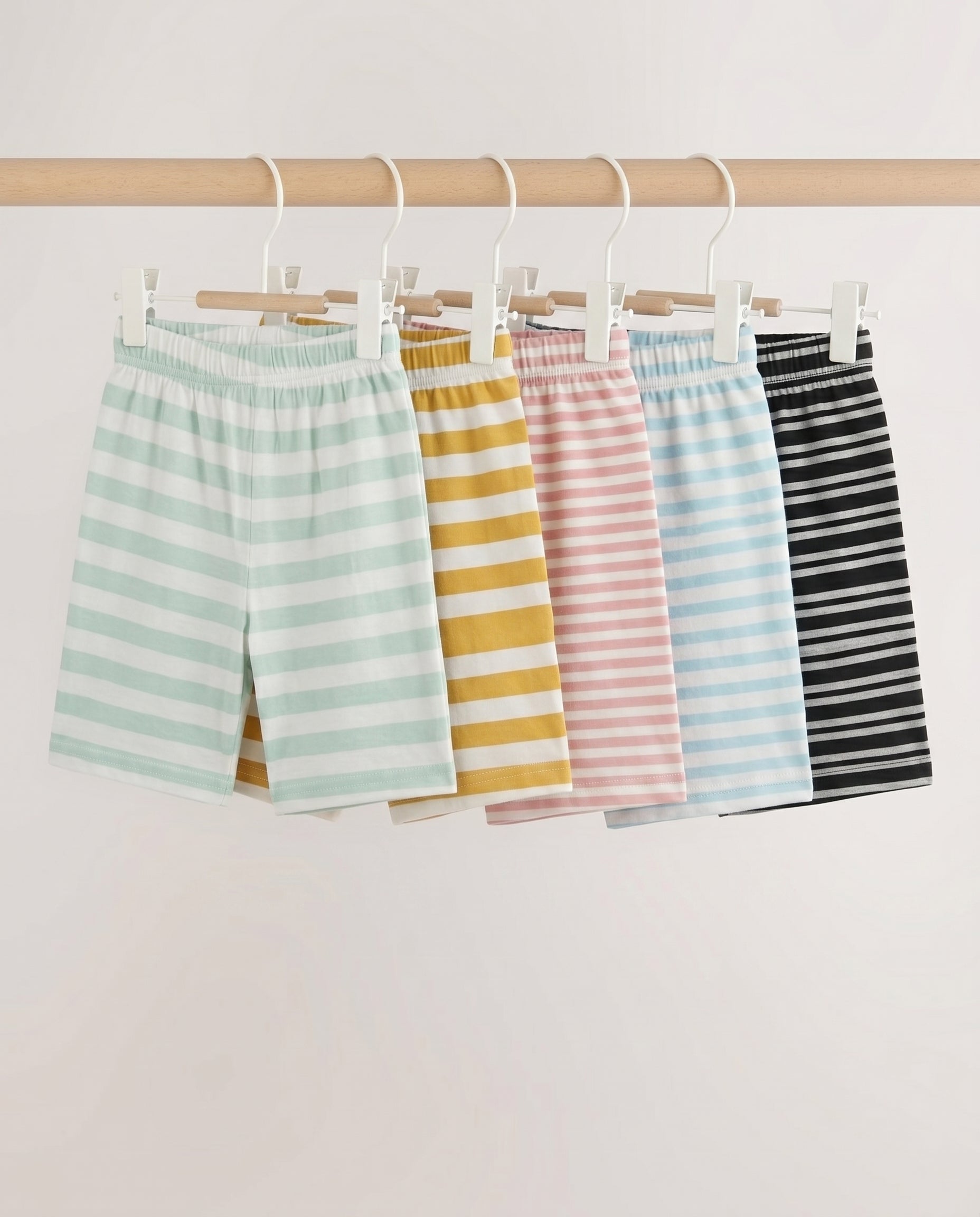 Five kids' striped pull-on shorts fanned out flat lay — mint, mustard, pink, blue, and black stripe patterns with elastic waistband, on light grey background.