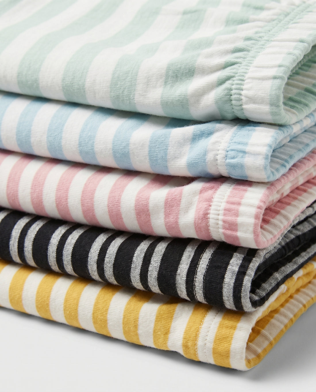 Five kids' striped pull-on shorts fanned out flat lay — mint, mustard, pink, blue, and black stripe patterns with elastic waistband, on light grey background.