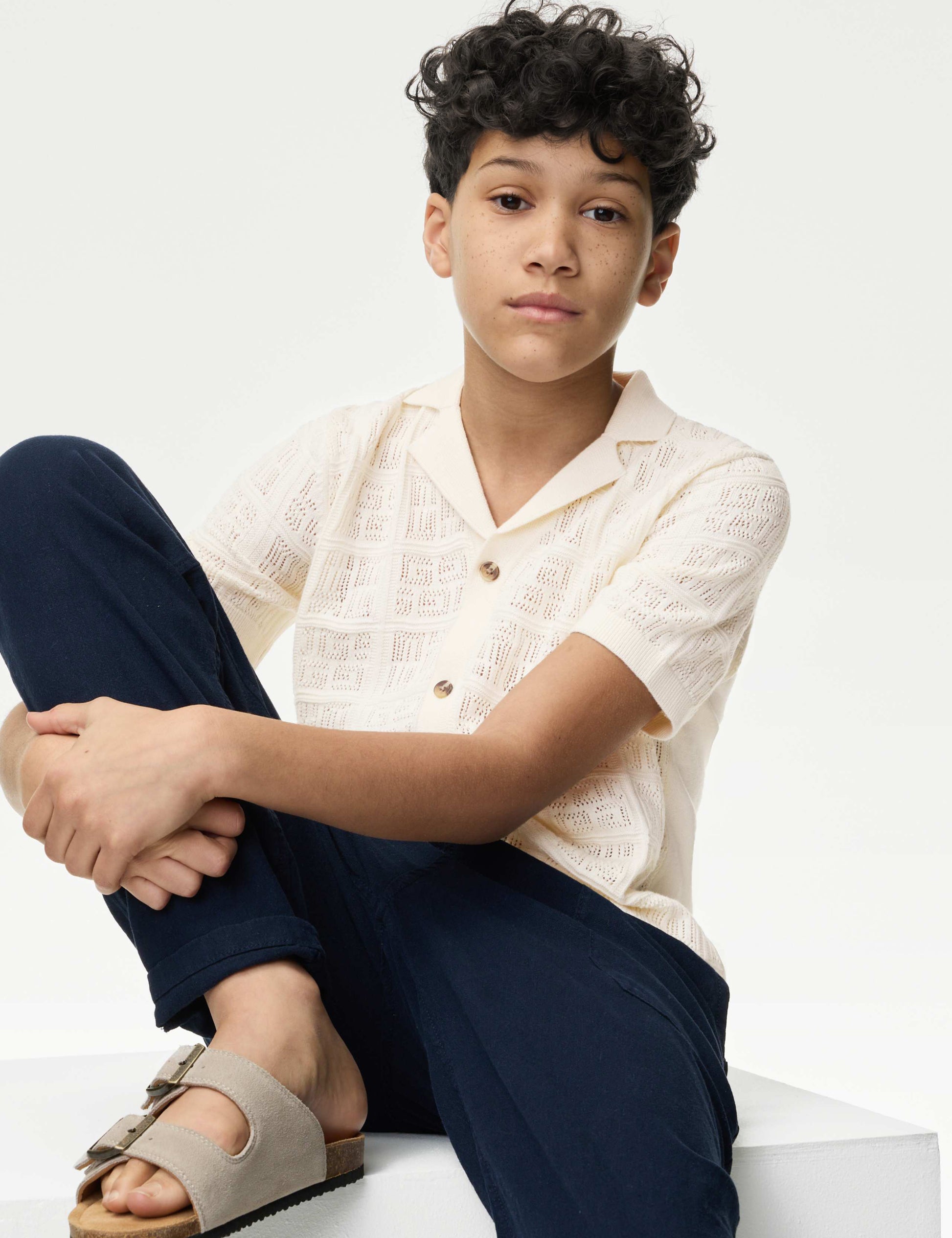 Young person wearing a white shirt and navy pants sitting on a white surface.