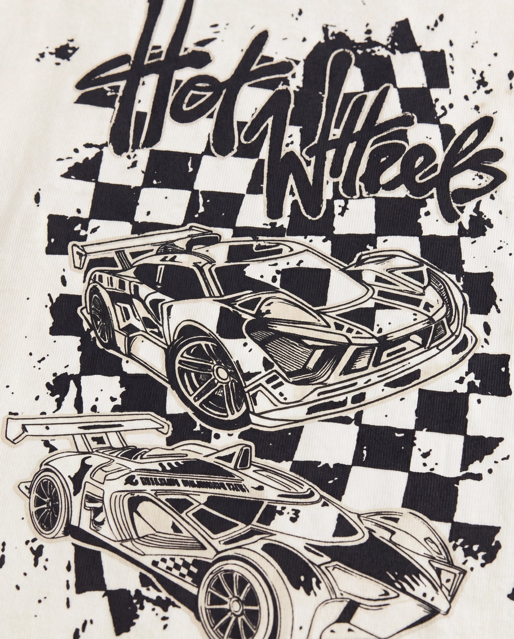 Close-up of the back graphic on an off-white toddler tee showing a black screen print of two Hot Wheels race cars over a splatter checkerboard background with graffiti-style "Hot Wheels" lettering.