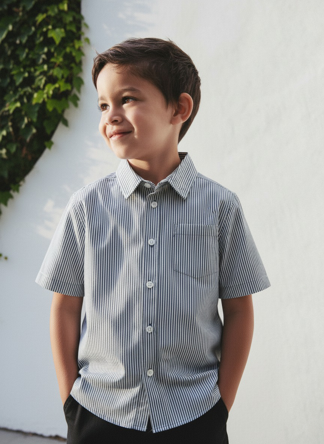 Young boy wearing a striped shirt standing against a white wall with greenery.