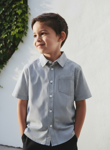 Young boy wearing a striped shirt standing against a white wall with greenery.
