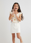 Young girl clapping and smiling in a white outfit with floral details on a light gray background