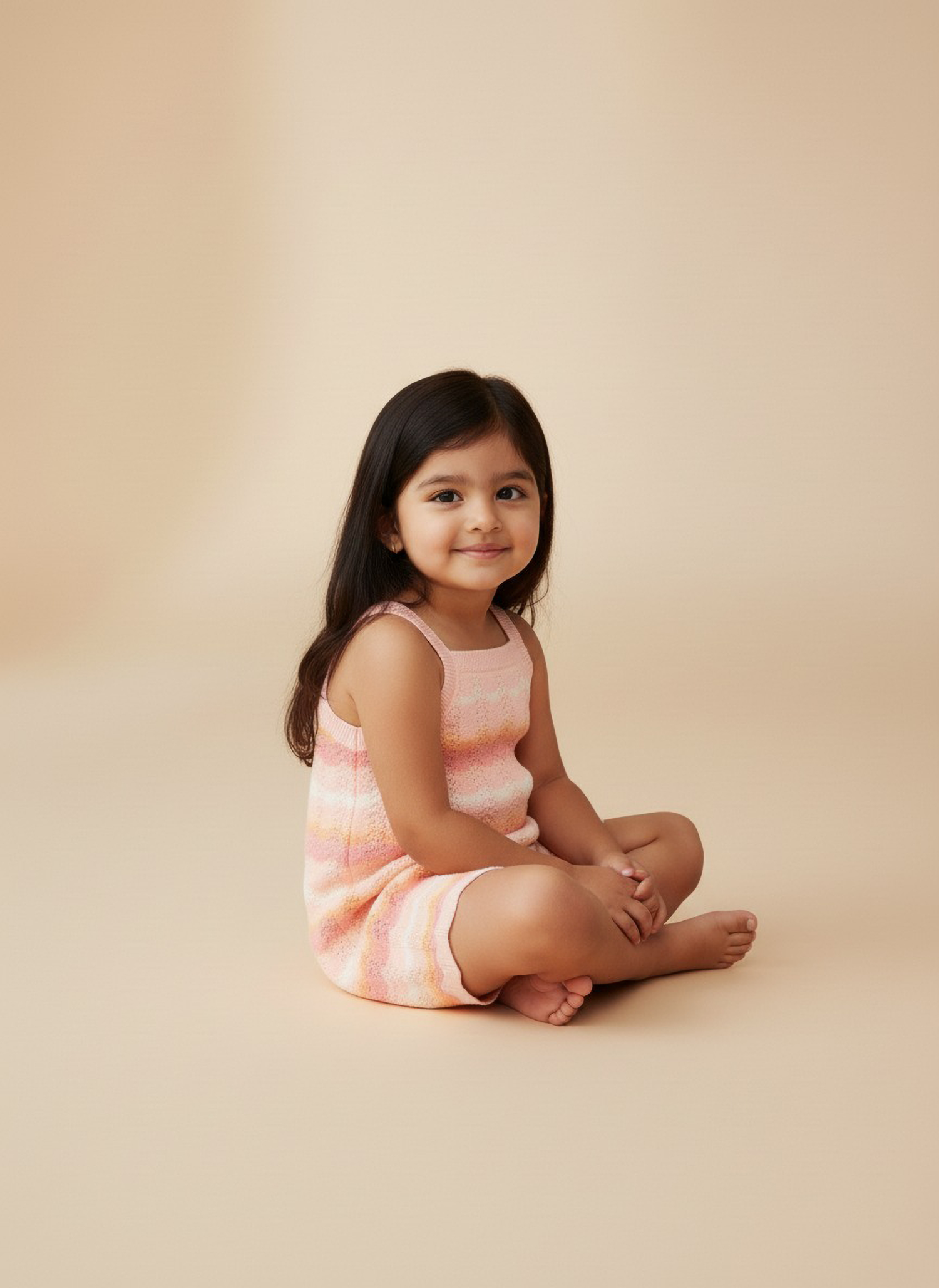 Young girl sitting with legs closed wearing a striped Jumpsuit against a beige background