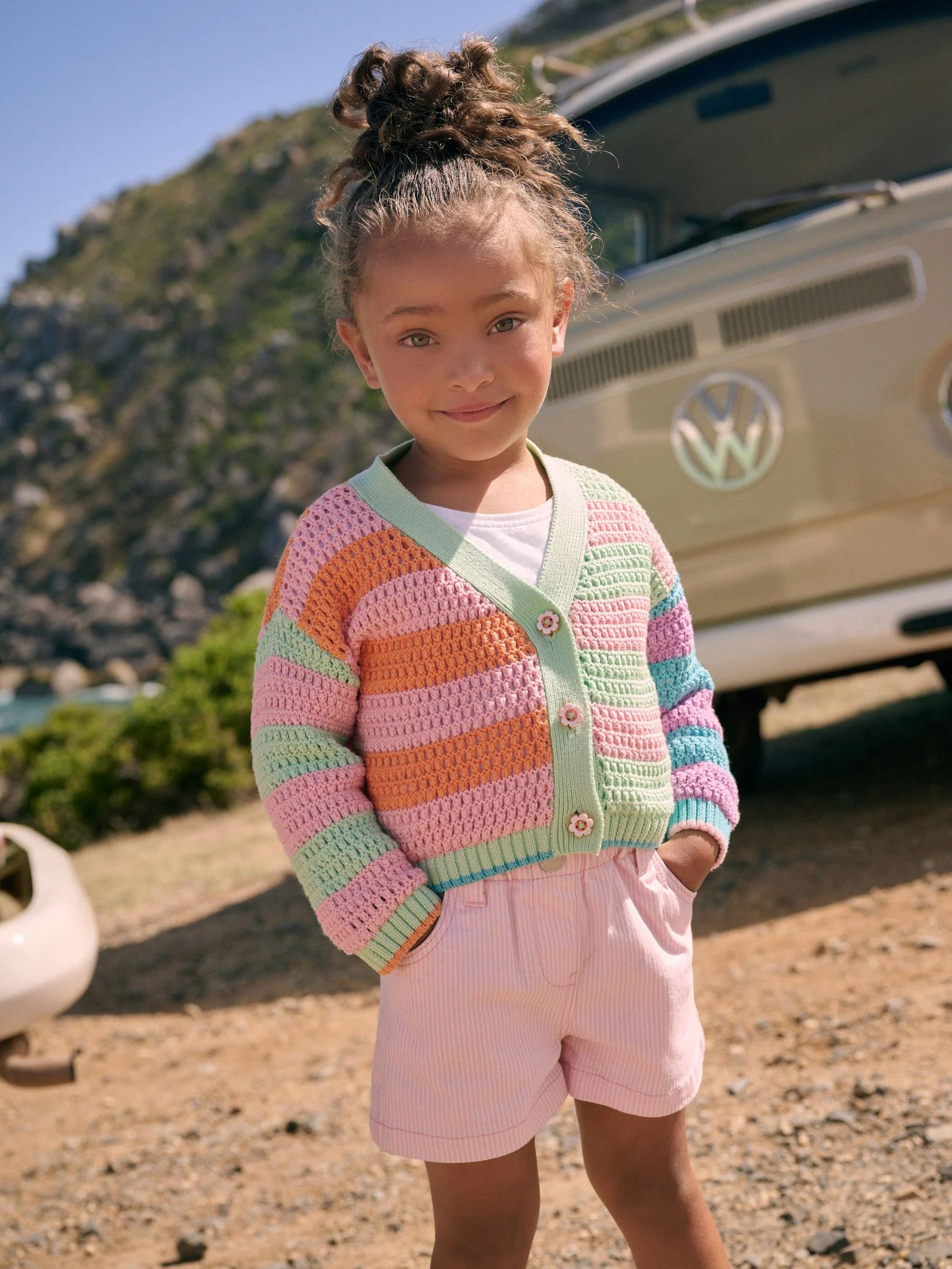 Young girl wearing a colorful striped sweater and pink shorts standing in front of a Volkswagen van.