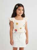 Young girl wearing a white crochet top with colorful patches and a white skirt on a light gray background