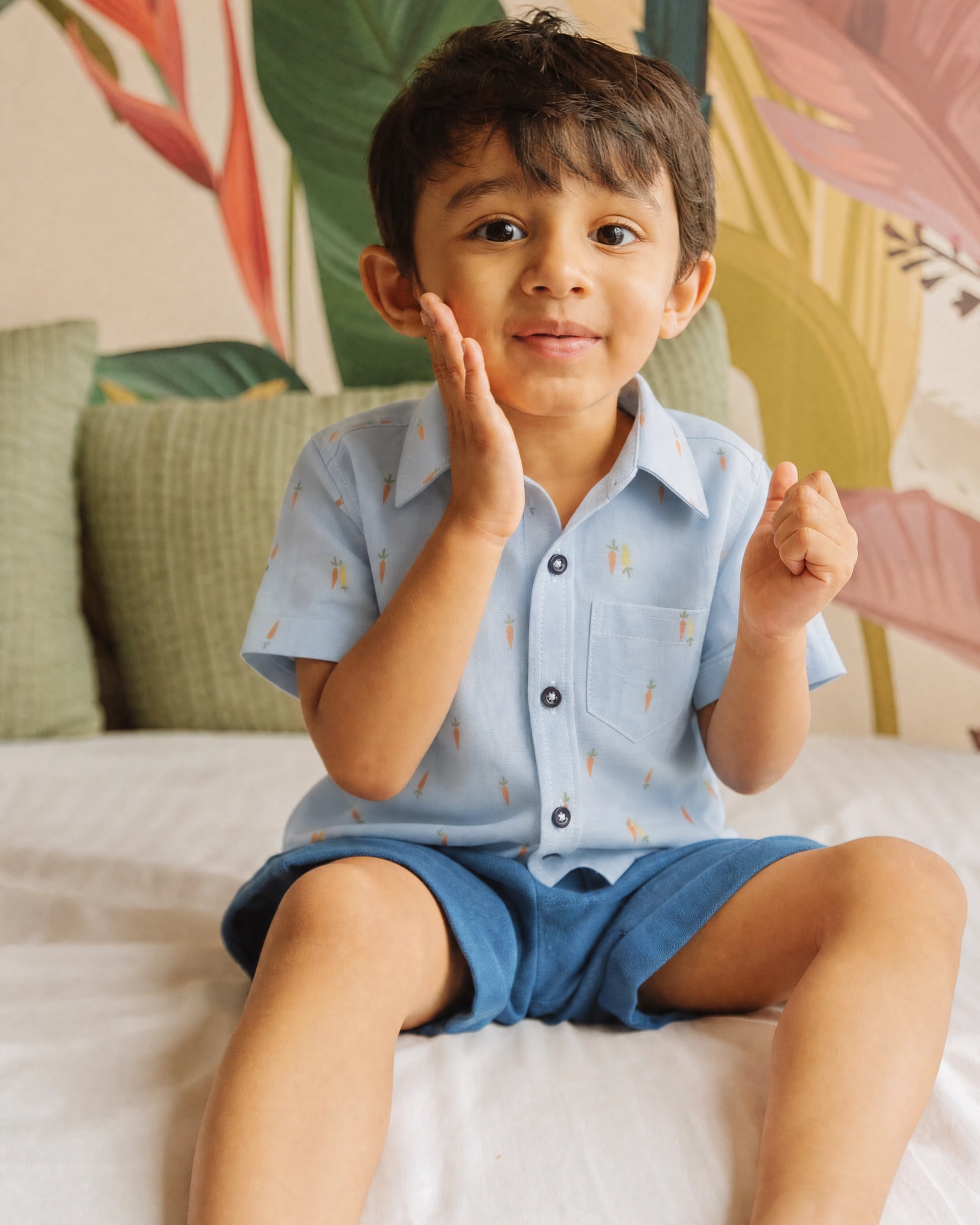 Boy sitting on a bed wearing a PTex Fashion sky blue carrot print cotton shirt with navy shorts