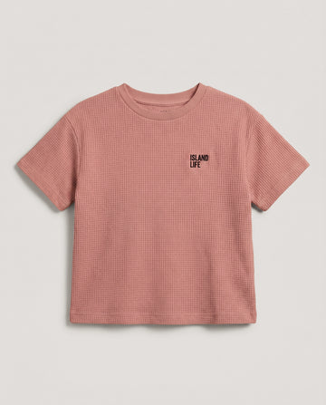 Boy's rusty rose waffle cotton t-shirt with "Island Life" black embroidery on the chest, relaxed boxy fit and ribbed crewneck, photographed flat on a soft grey backdrop.