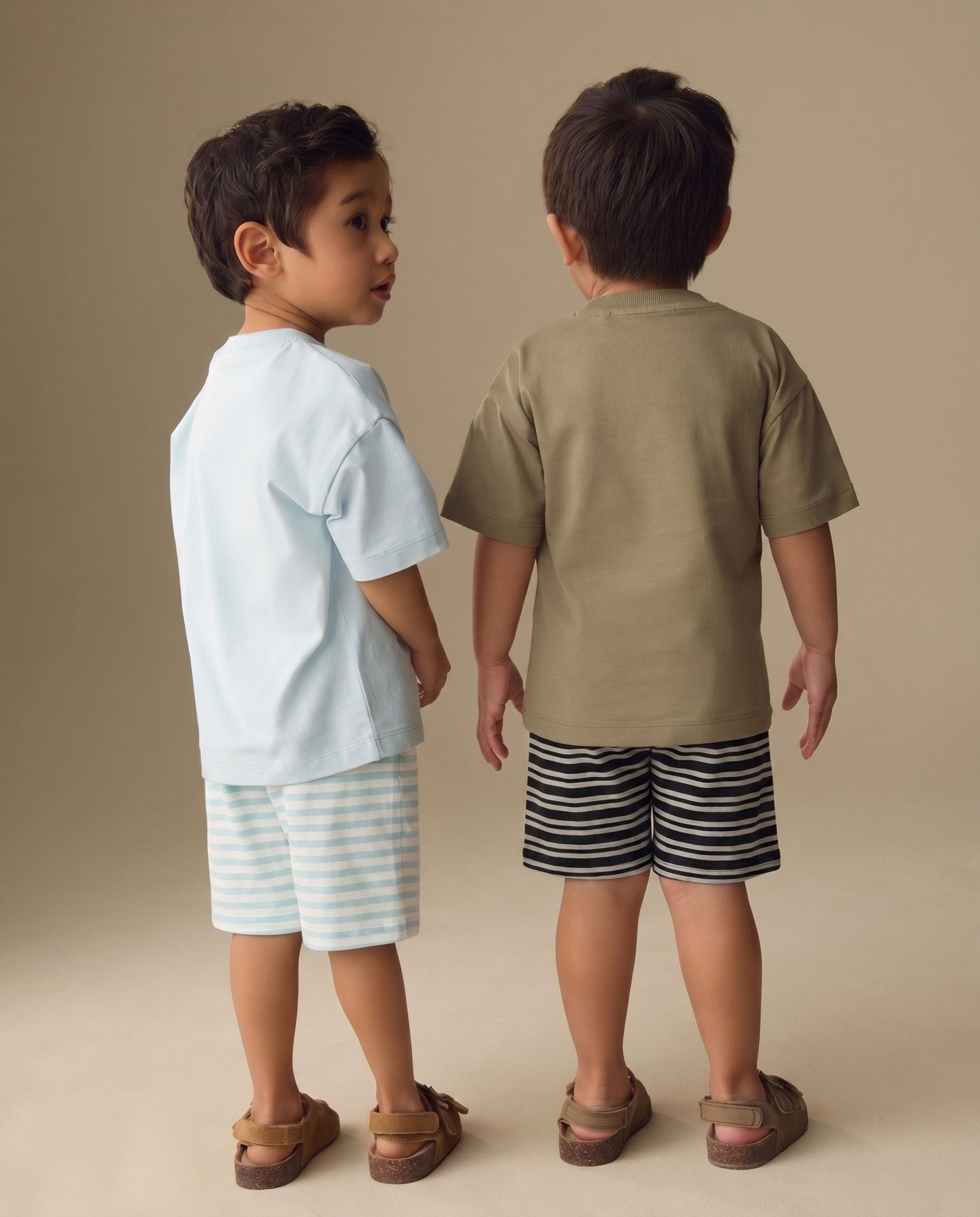 Two children standing back-to-back wearing light blue and beige shirts with striped shorts on a beige background from PTex Fashion