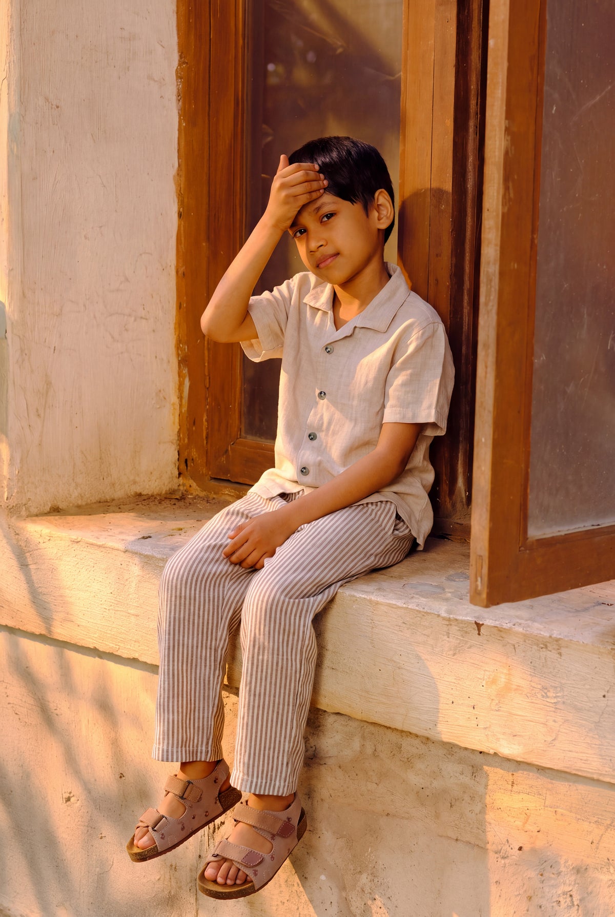 A young boy sitting on a stone ledge next to a wooden door, wearing a light beige short-sleeve button-up shirt, striped trousers, and brown sandals.