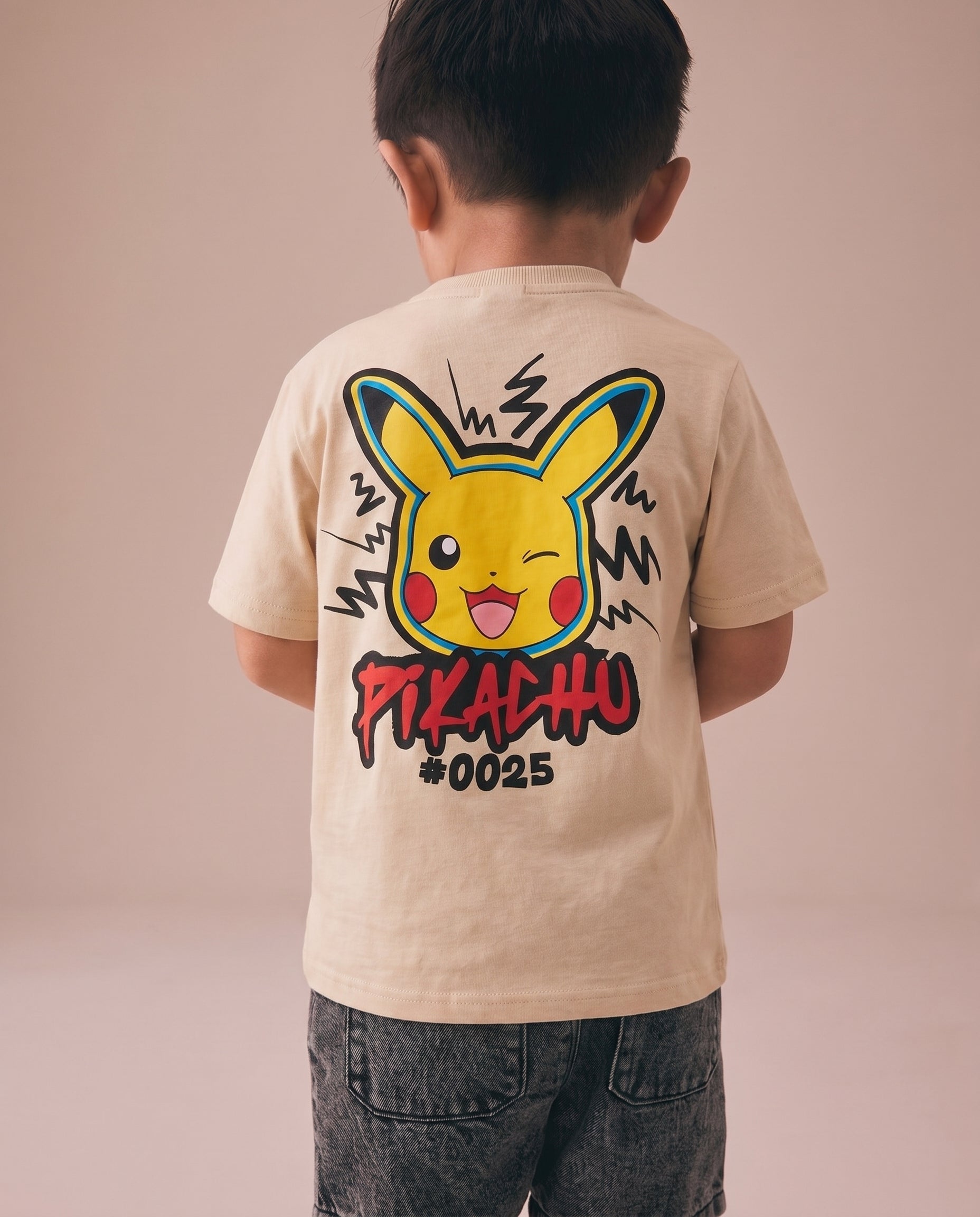 Young boy showing the back of a cream kids' t-shirt with a bold graffiti-style Pikachu winking face graphic and lightning bolt details