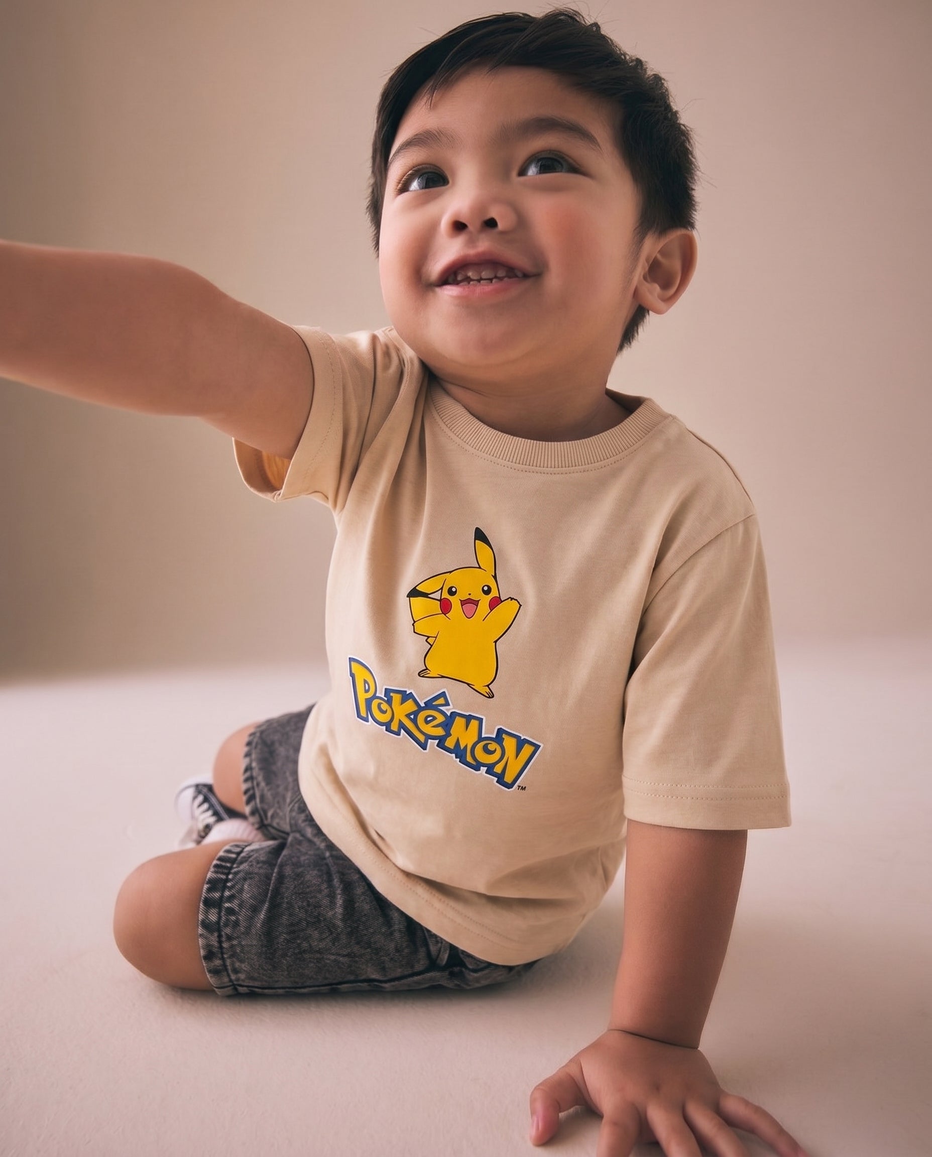 Young boy wearing a cream kids' t-shirt with a cheerful Pikachu full-body graphic print and cartoon logo on the front