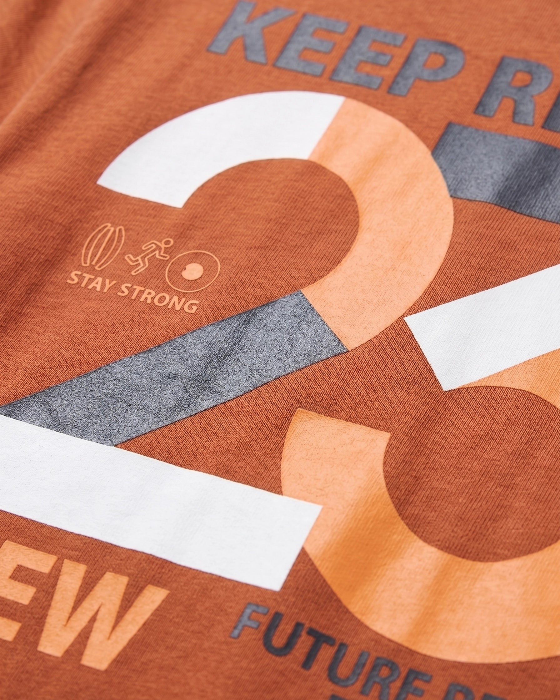 Close-up detail of bold "23" graphic print on rust orange boys' t-shirt featuring orange, white, and charcoal color blocks with "Stay Strong" icon