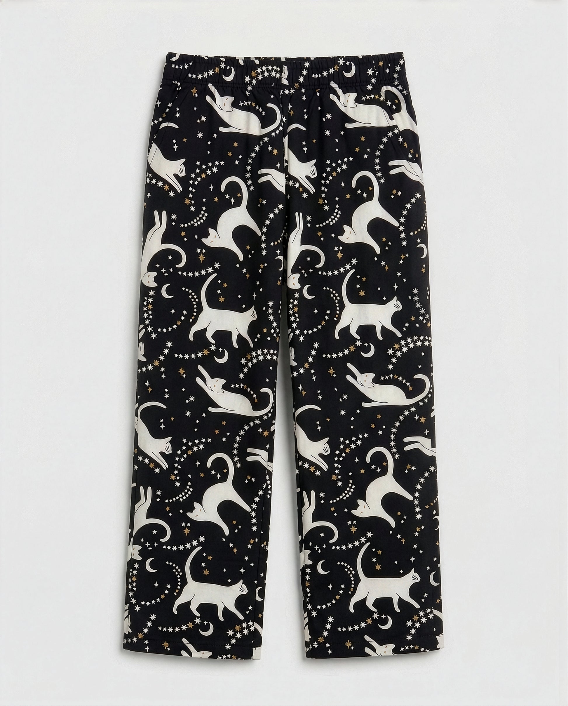 Girls' straight-cut wide-leg palazzo pants in deep black woven cotton with all-over celestial cat print — ivory white cat silhouettes in playful poses surrounded by gold and white star clusters and crescent moons, featuring full elastic waistband, flat-lay product photography on white background