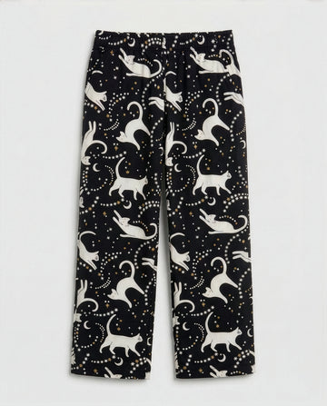 Girls' straight-cut wide-leg palazzo pants in deep black woven cotton with all-over celestial cat print — ivory white cat silhouettes in playful poses surrounded by gold and white star clusters and crescent moons, featuring full elastic waistband, flat-lay product photography on white background