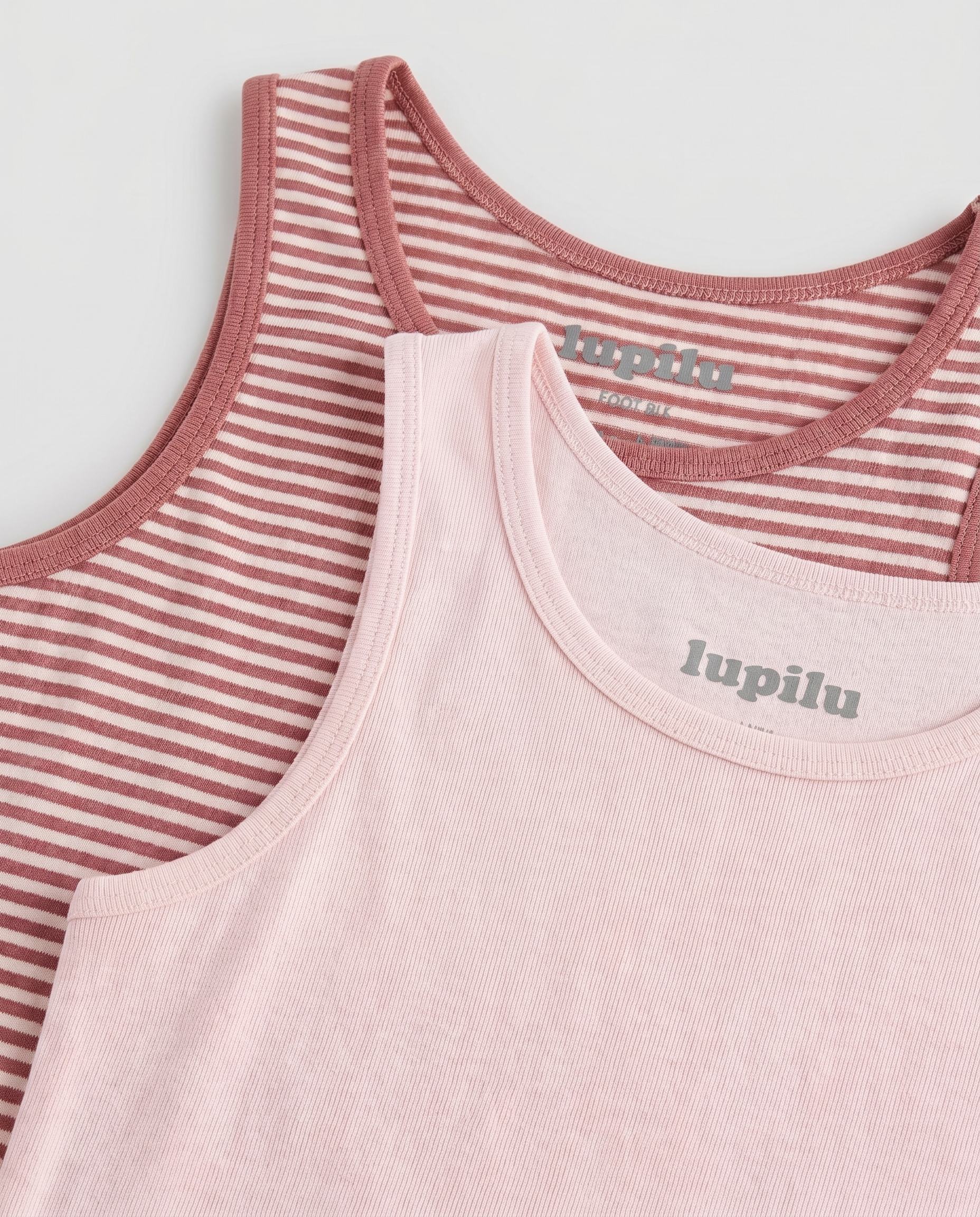 Close-up flat lay of Lupilu girls' two-pack tank top set showing neckline detail, with solid blush pink ribbed tank layered over a rose and cream horizontal striped tank, both featuring bound necklines and tonal Lupilu logo prints.