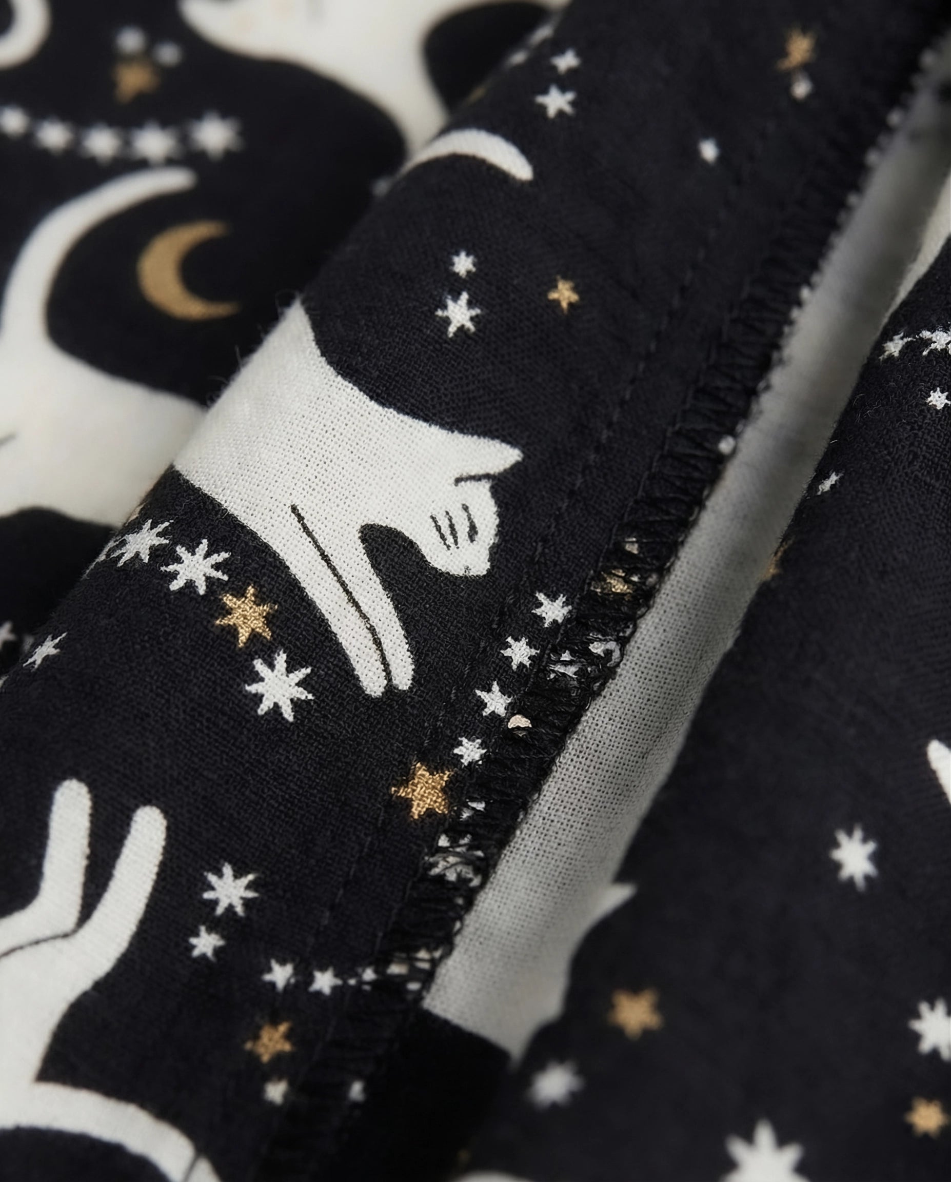 Extreme close-up of girls' black woven cotton palazzo pants showing inner leg seam with serged overlock stitching finish, fabric texture detail, and celestial cat print with white cat silhouettes, gold stars, and crescent moon motifs on black cotton