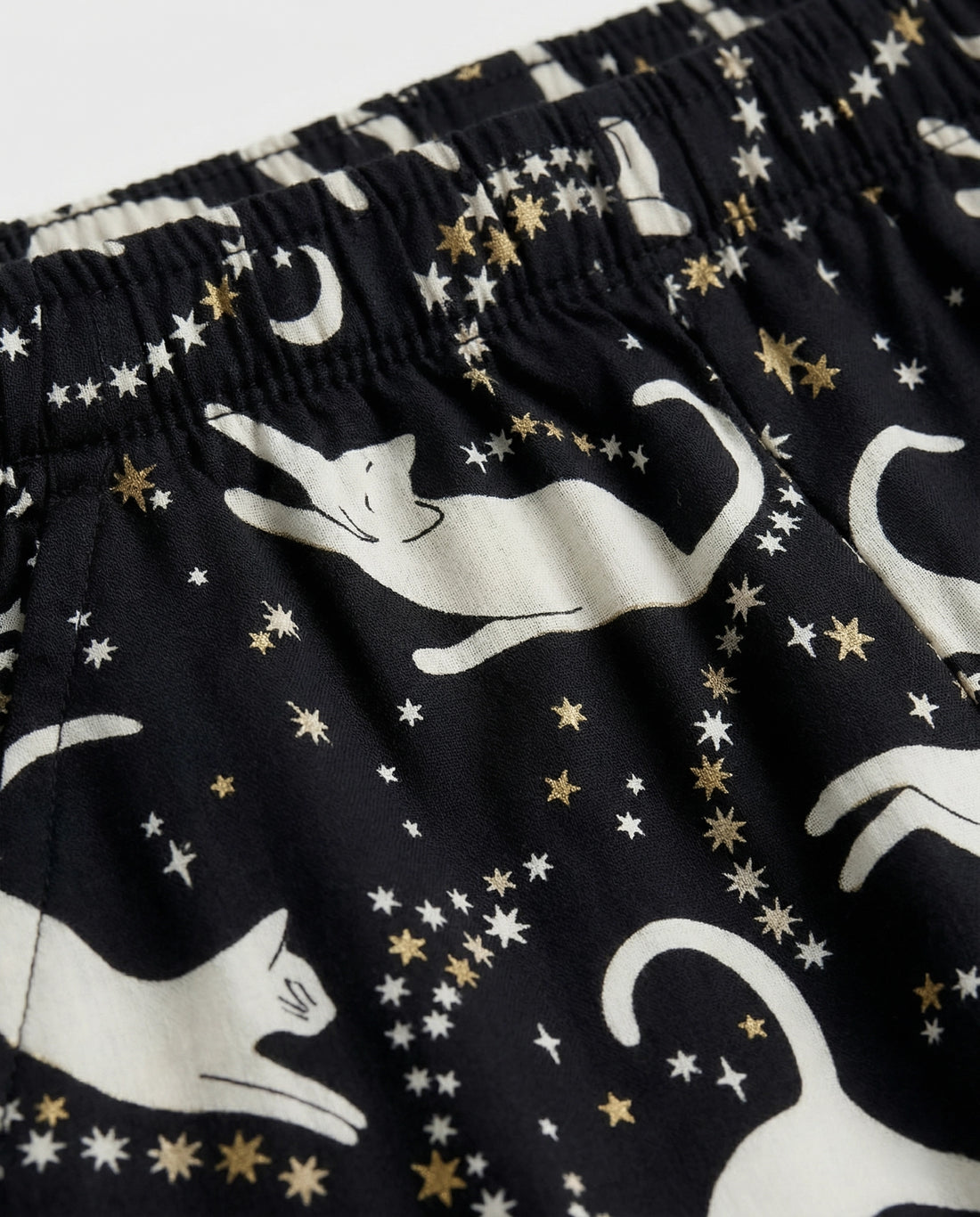 Girls' straight-cut wide-leg palazzo pants in deep black woven cotton with all-over celestial cat print — ivory white cat silhouettes in playful poses surrounded by gold and white star clusters and crescent moons, featuring full elastic waistband, flat-lay product photography on white background